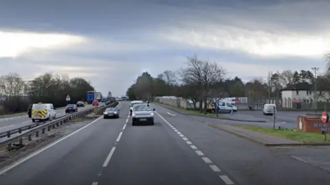 A dual carriageway with traffic driving on both sides and a residential housing to the right of the road