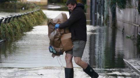 A man in wellington boots carries cardboard boxes through floodwater