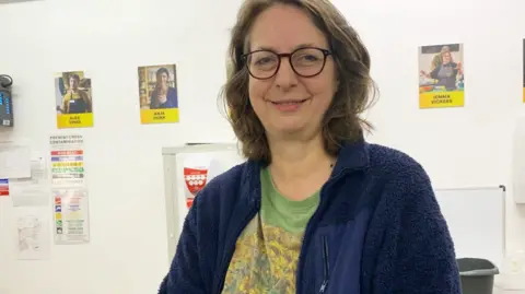 Nicky Whitfield smiles at the camera and behind her is a light wall full of other people's pictures and posters