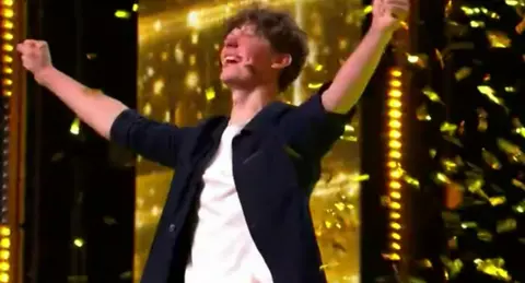 Tom Dymond/Talkback Thames A beaming BGT golden buzzer winner Rafferty Coope has both hands high in the air and his eyes are closed in disbelief as he celebrates his golden buzzer moment. Wearing a white T-shirt under his open black shirt his sleeves are rolled up to the elbows as golden confetti falls from the roof of the building all around him. 