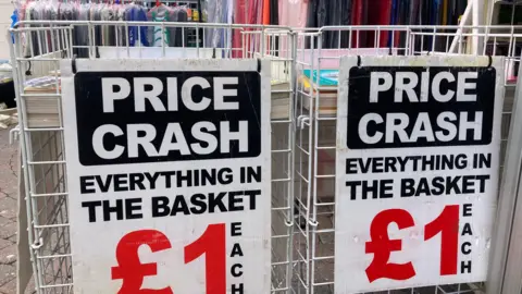 A shot of Worcester market, showing two baskets with signs on saying 'price crash, everything in the basket is £1 each'. You can also see hangers with clothes on them.