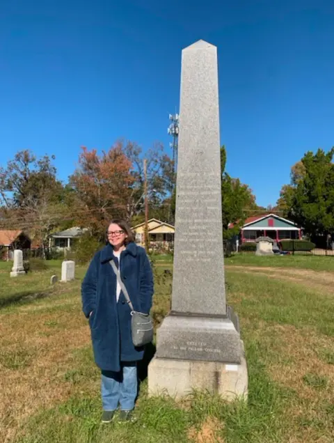 Lydia Melton Lydia Melton pictured next to a tall grey obelisk. The monument is surrounded by grass and houses are visible in the background. Lydia has dark long hair and is wearing glasses. She is wearing a long, warm coat and has a bag over her shoulder. 