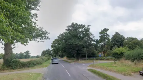 A silver car drives away from along a road, bordered by grass verges, trees and fields. There is a crossroads. 