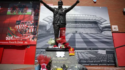 EPA A statue of Bill Shankly on a stone podium is surrounded by flowers and scarves, with the stadium in the backdrop