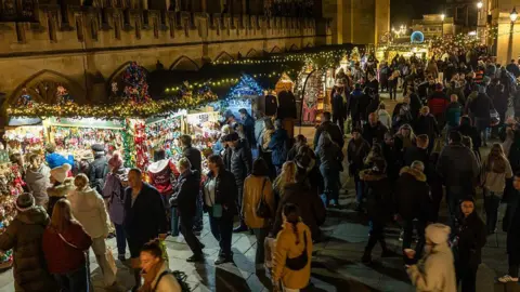Getty Images Large crowds are walking past Christmas market stalls in the dark. The stalls are brightly lit with Christmas lights and sit in front of a historic Bath stone building.