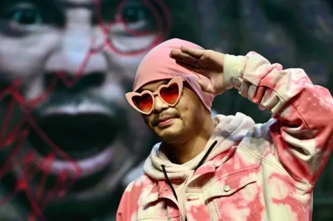 Namewee wearing a pink beanie, pink heart-shaped sunglasses and a pink denim jacket, raising his hand to his forehead in a saluting pose.