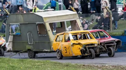 Lime Jelly Photography Beat up three-wheeled cars and a green caravan are driving around a track with a small crowd behind on a grassy bank.