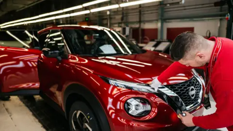 An employee, wearing a red shirt, inspects a red sports utility vehicle on the production line with Nissan Juke and Nissan Qashqai at the Nissan factory in Sunderland, UK, on Friday, 24 November, 2023.