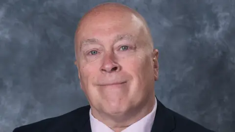 A bald man is wearing a pink shirt and a black blazer. He is smiling at the camera. 