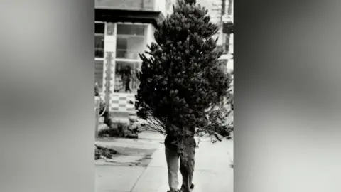 Getty Images A man walks down the street with a Christmas tree, which obscures the top half of his body.