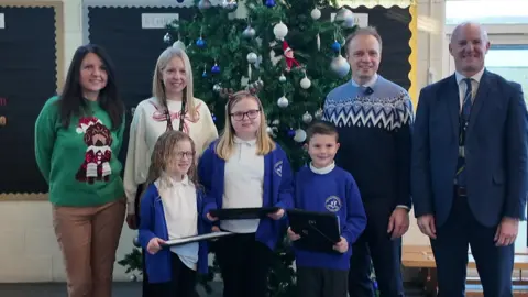 Two female teachers dressed in christmas jumpers, stand enxt to two men in blue christmas jumpers. In front of them are three children in school uniforms holding certificates. All are smiling at the camera..