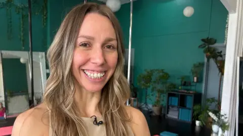 BBC is standing inside a brightly lit fitness studio with teal-colored walls. The space features large mirrors, hanging plants, and white spherical light fixtures. In the background, there are potted plants and a shelving unit with storage boxes. A microphone is clipped to the person’s beige top, and a vertical pole is visible behind them, indicating the studio is used for pole fitness.