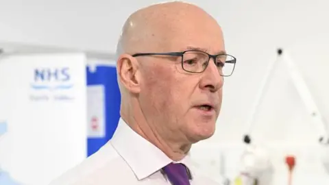 PA Media First Minister John Swinney lasting  successful  beforehand   of an NHS distant   vale  sign. He is bald and wearing glasses. He has a achromatic  garment  connected  and a purple tie.