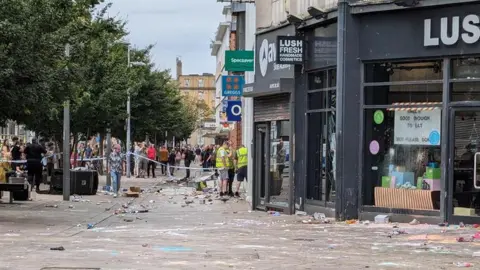 The outside of a number of damaged shops, including Lush and O2, with broken windows and stolen items on the pavement. The area is cordoned off and workers in hi-vis vests are assessing the damage, watched by a crowd of people standing behind the tape in the distance. 