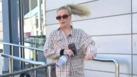 BBC A woman with long blonde hair tied up in a ponytail, with a beige checked shirt and sunglasses, leaves court. She has a water bottle and a mobile phone.