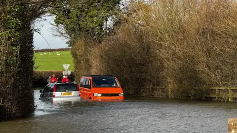 Dorset Council Emergency services attending a rural area in Dorset where severe flooding has almost submerged two vehicles. Sheep can be seen grazing on a meadow in the distance.