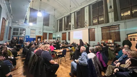 Large town hall room packed with people sat behind tables