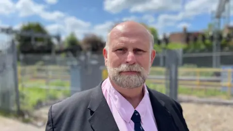 A picture of the Mayor of Bedford Tom Wootton. He has a grey and white beard and moustache and is wearing a pink shirt and a black jacket and tie. He is stood in front of a railway line although the background is out of focus.