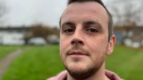 Marcus Johnson stands in front of a grassed area with cars in the background.