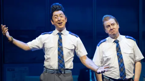 Two performers in matching airline-style uniforms perform on stage in The High Life: The Musical, gesturing as they sing under blue stage lighting.
