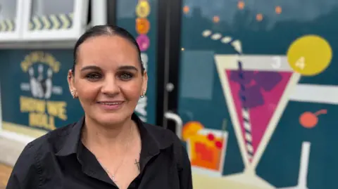 Michelle Hannigan. She has dark hair tied back, wearing small hoop earrings and a black shirt.