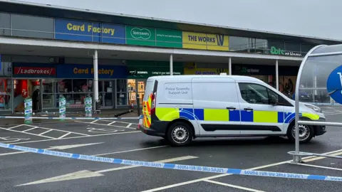 A white transit van with police scientific support unit on its back side panel and yellow and blue markings parked up in a car park behind blue and white police tape. Behind it is a single-storey row of shops under a low-lying roof, supported by pillars. Stepped back behind it are the shops, including a Ladbrokes with a destroyed door on its hinges, with its glass shattered on the ground. 