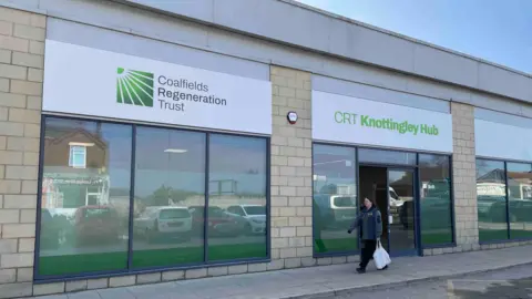 BBC/Spencer Stokes The outside of a bricked building with a person walking past. There is a white sign on the buiding with green writing on which says 'Coalfields Regeneration Trust CRT Knottingley Hub. 