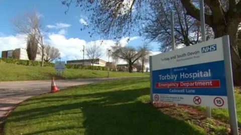 A blue and white sign on the grass outside Torbay Hospital welcoming visitors to the site. The sign is for the emergency department. It is on a grass bank with a traffic cone to the left of it. There is a road leading to the hospital. 