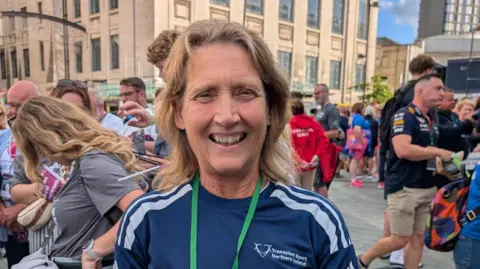 Team NI Kay Hack has long blonde hair and is wearing a navy T-shirt and green lanyard. She is standing in front of a large crowd of people. 