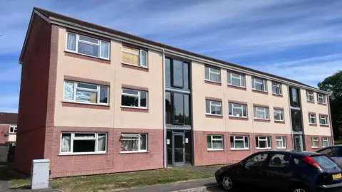 Picture shows a three storey block of flats, with cars parked in front. An upstairs  window is boarded up. 