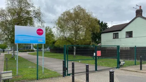 Phil Shepka/BBC A view of the All Saints Academy from outside the school gates. A bright blue sign includes the schools circular red logo. The gates are green. There is a tree lined path which obscures the school in the background.