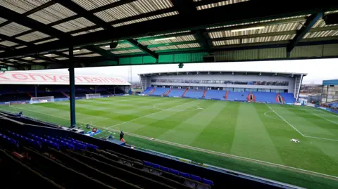 A football ground can be seen from the top of a stand looking out on to the pitch, with three other stands seen either side of the grass. The seats are blue. The pitch is empty spare a man waking near the dug. 