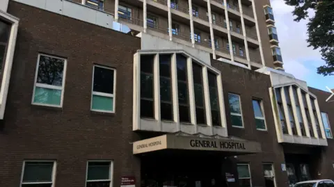 A photo of the front entrance of Jersey General Hospital. It is a large brown brick building. 