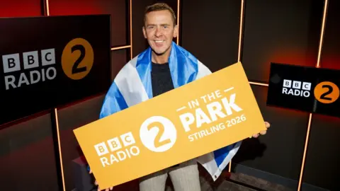 BBC Scott Mills, wearing a Saltire flag and holding a Radio 2 In the Park Stirling 2026, in a radio studio
