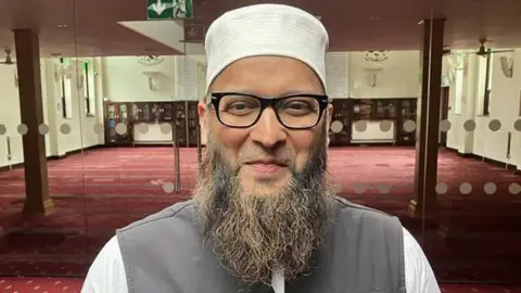 A man with a large beard, black glasses and a white hat. He is stood inside a mosque with glass and red carpet behind him.