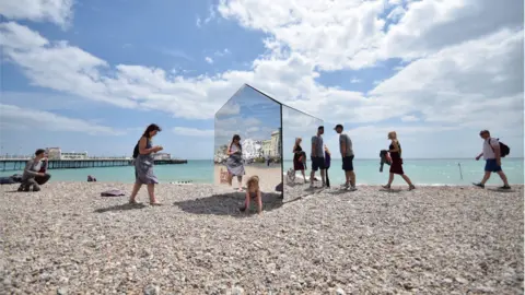 Mark Sephton On a sunny day, people stand on a pebbled beach looking at a mirrored beach hut, which sits near the sea. The sea in the distance is light blue and the sky is blue with some clouds.
