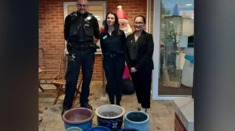 A male police officer stands next to two women behind some plant pots