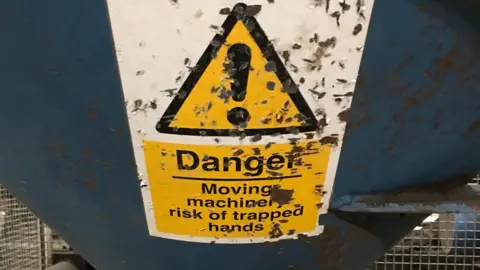 HSE A worn-out warning sign on a piece of machinery that has a black exclamation mark in a yellow triangle with a black edge. It reads Danger. Moving machinery - risk of trapped hands