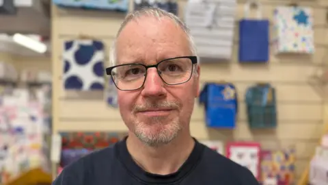Stephen stands in front of a wall holding wrapping paper and bags. He wears dark glasses and has a grey goatee beard.