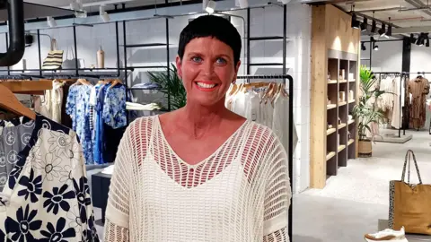 A woman is standing in a shop that sells women's clothes and bags. She has short, black hair tightly cropped to her face and is wearing a white vest covered by a mesh white top.