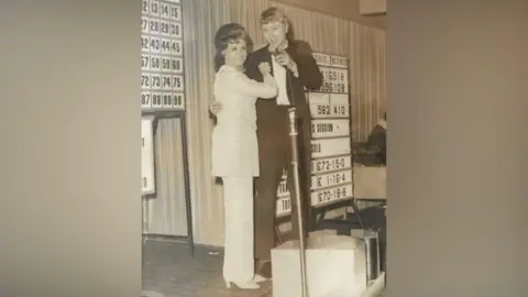 James Dawn A black and white photo of James as a young man on stage with his arm around a young woman who is dressed in a white trouser suit. He is wearing black tie and holding a mic and there are boards with bingo numbers in the background. 