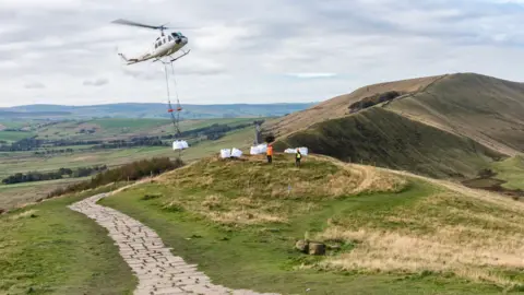 A helicopter carrying large white bags of stones over a grassy part of the countryside