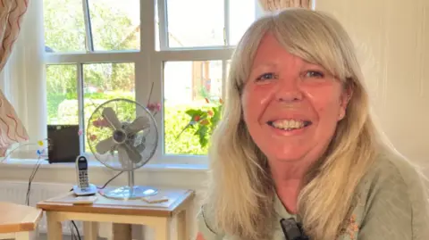 Karen MacDonald sits in her living room and smiles at the camera. There is a window and a fan behind her. She has long, blonde hair with a fringe.
