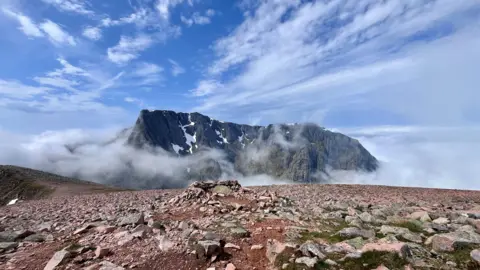 Susan Morrison A dramatic mountain landscape showing rugged, rocky peaks rising through low cloud and mist, with patches of lingering snow beneath a bright blue sky streaked with thin clouds.