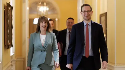No 10 Downing Street Rachel Reeves and Darren Jones walk down an ornate interior yellow-painted corridor. Reeves on the left of the photo, is in a light teal business suit wit a white top underneath her jacket. Beside her, on the right of the picture, Jones is in a dark blue suit with a light blue shirt and red tie.