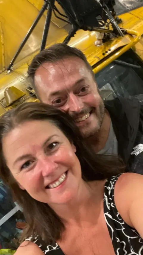 Anna-Louise Stubbings A selfie of Anna-Louise says Zach who are standing in front of a Sea King helicopter. Anne is wearing a sleeveless black and white dress and has long dark hair. Zach is in dark overalls.