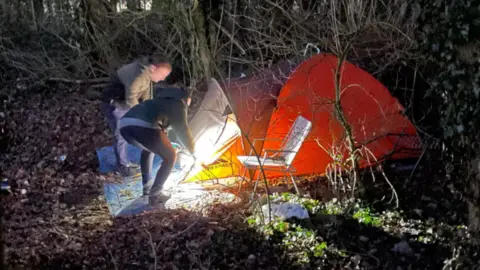 Two street outreach workers use a torch to check an orange tent at night