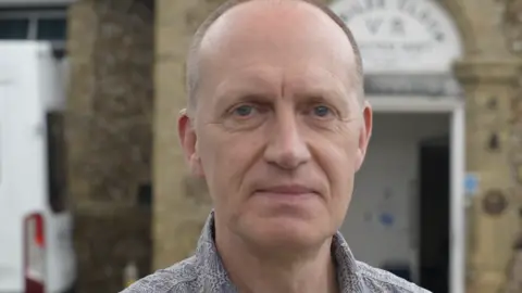 Jamie Niblock/BBC A man who is largely bald with some short hair around the side of his head. He is slightly smiling at the camera. He wears a grey shirt with some patterns on it. He is standing outside, in front of a village hall.