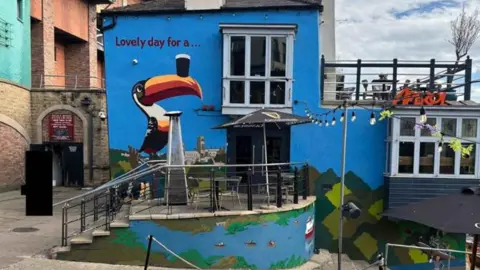 The Boat Club mural shows a large toucan balancing a pint on its beak next to the venue's entrance door. Durham Cathedral and the River Wear are pictured in the background. Most of the wall is blue. "Lovely day for a..." is written above the toucan. The building has a glassed terrace on its lower floor and an open one at the top. There is a bright orange Aperol sign on the roof of the indoor terrace.