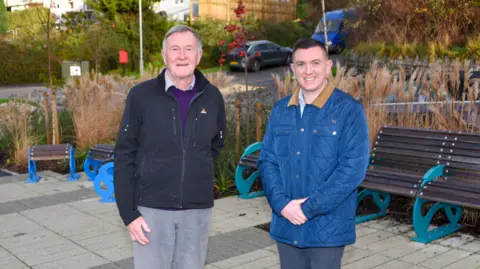 Cornwall Council Alan Stanhope and Kieren Couch standing by one another. Alan has short grey hair and is wearing a dark jacket and grey trousers. Kieren has short dark hair and is wearing a blue jacket with a brown collar. He is wearing dark trousers. They are standing in front of some of the route's new benches.